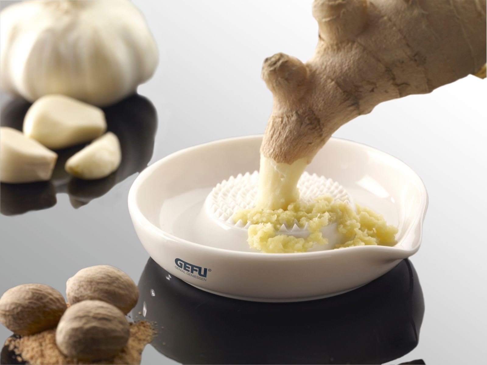 Runde Ingwerreibe aus Keramik mit geriebenem Ingwer, von oben fotografiert. Im Hintergrund sind Knoblauchzehen und Muskatnüsse zu sehen.