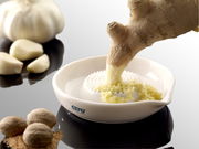 Runde Ingwerreibe aus Keramik mit geriebenem Ingwer, von oben fotografiert. Im Hintergrund sind Knoblauchzehen und Muskatnüsse zu sehen.