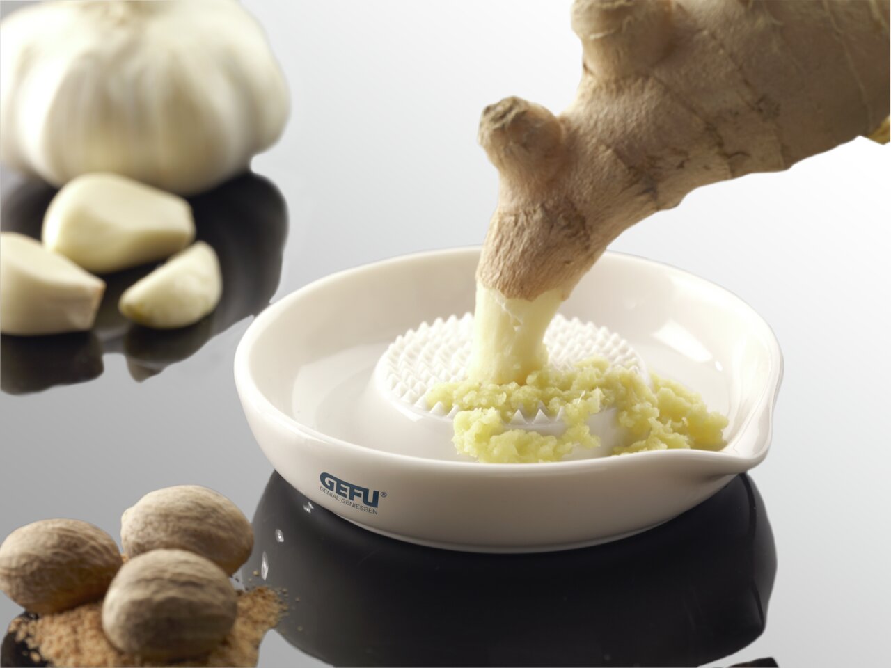 Runde Ingwerreibe aus Keramik mit geriebenem Ingwer, von oben fotografiert. Im Hintergrund sind Knoblauchzehen und Muskatnüsse zu sehen.