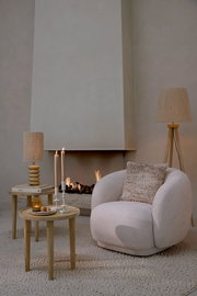 Gemütlicher, cremefarbener Sessel mit rundem Design, dekoriert mit einem strukturierten Kissen, vor einem modernen Kamin. Perspektive von vorne.