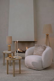 Gemütlicher, cremefarbener Sessel mit rundem Design, dekoriert mit einem strukturierten Kissen, vor einem modernen Kamin. Perspektive von vorne.