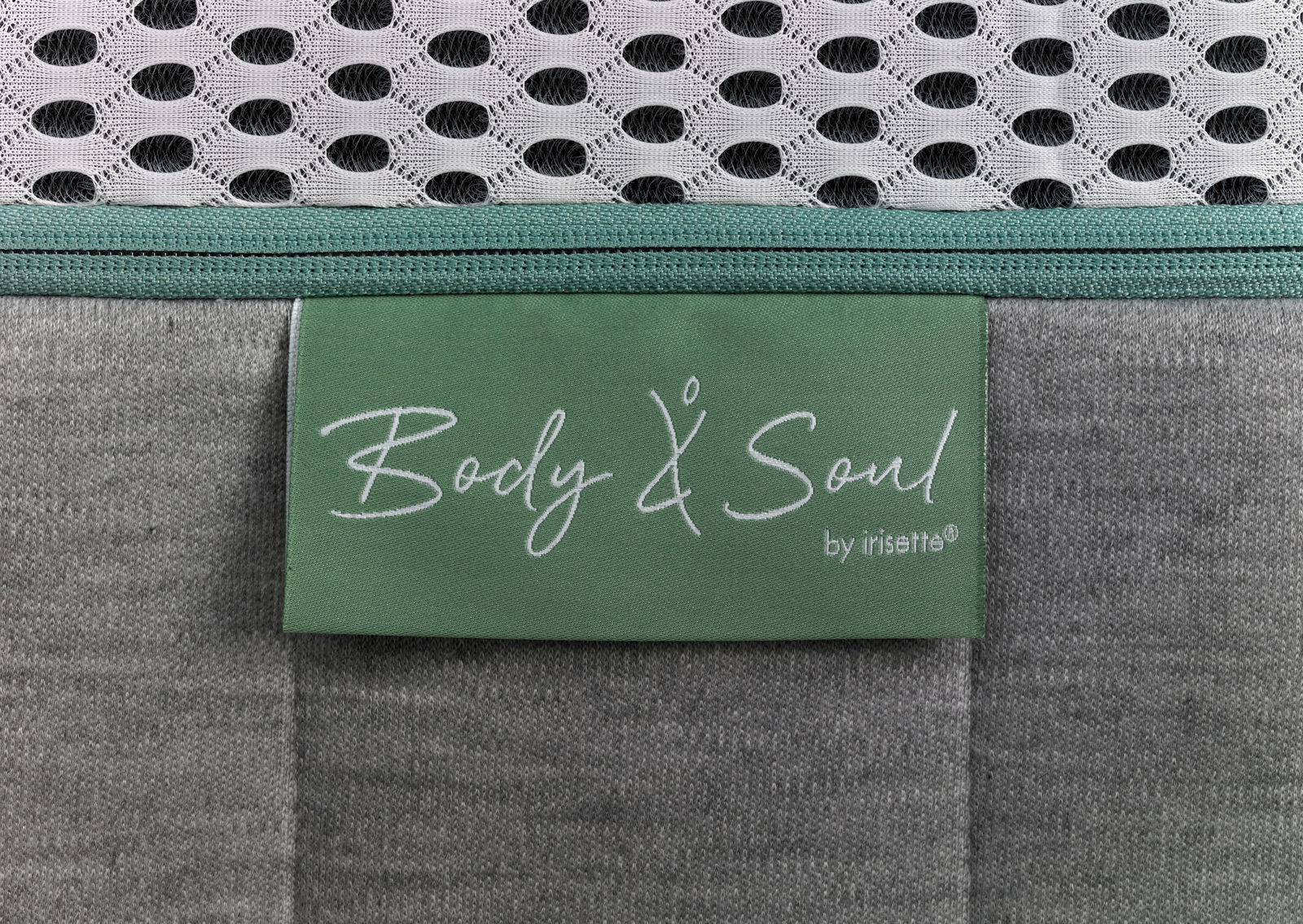 Nahaufnahme einer Matratze mit grünem Etikett 'Body & Soul by irisette', grauer Stoff und weißem Netzgewebe.