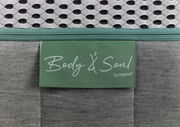Nahaufnahme einer Matratze mit grünem Etikett 'Body & Soul by irisette', grauer Stoff und weißem Netzgewebe.