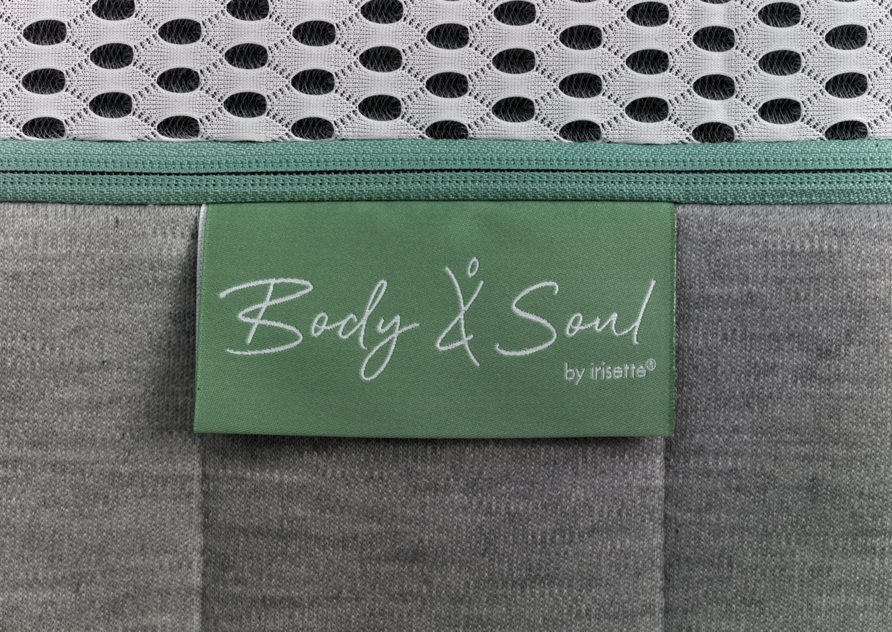 Nahaufnahme einer Matratze mit grünem Etikett 'Body & Soul by irisette', grauer Stoff und weißem Netzgewebe.