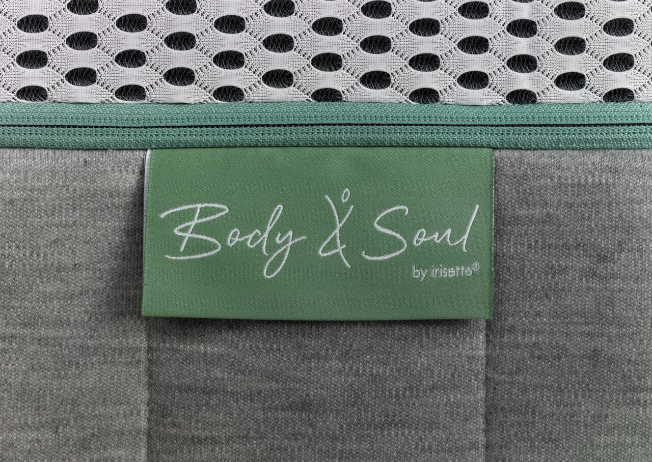 Nahaufnahme einer Matratze mit grünem Etikett 'Body & Soul by irisette', grauer Stoff und weißem Netzgewebe.