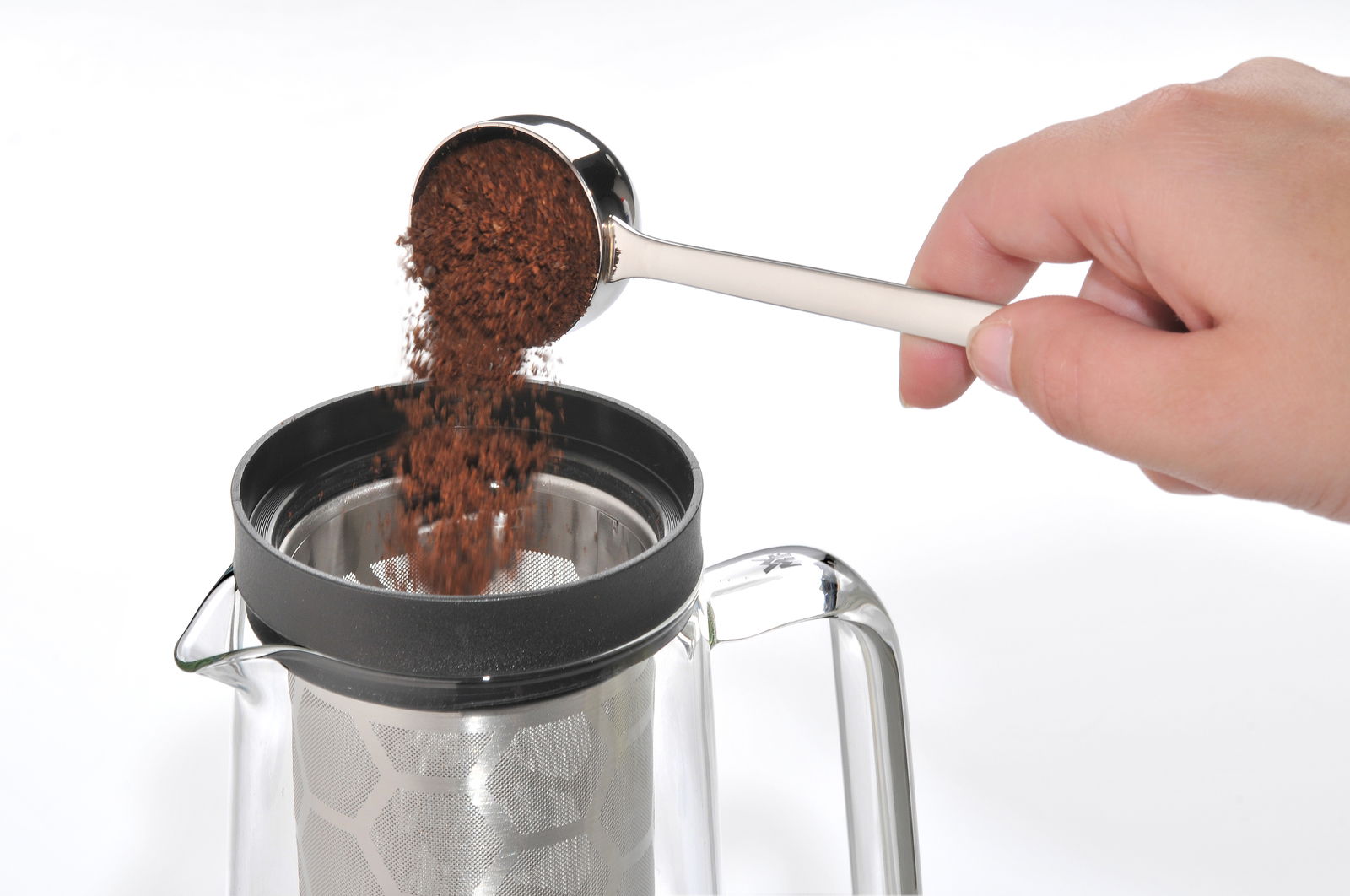 Hand hält einen Kaffeemaßlöffel über einer Kaffeekanne und schüttet gemahlenen Kaffee hinein, seitliche Perspektive.