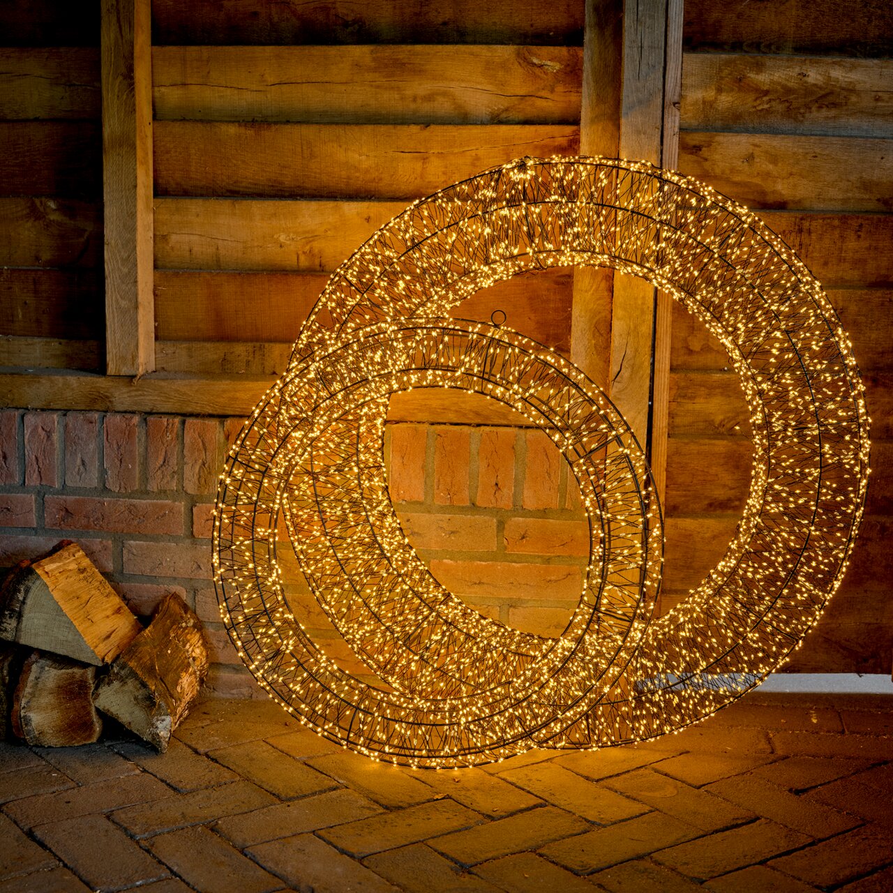 Lumineo LED-Kranz X-MAS Ø58cm mit 1440 LED``s Zwei leuchtende LED-Kränze in einer rustikalen Umgebung, frontal betrachtet.