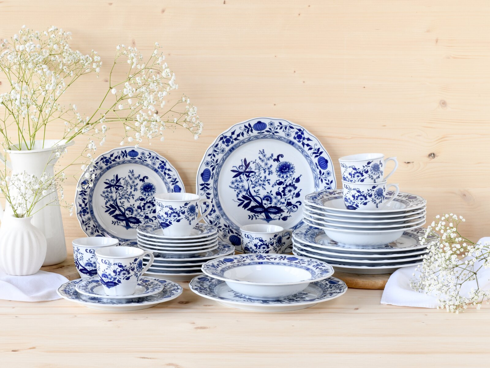 Blau-weißes Porzellan-Geschirrset mit floralem Zwiebelmuster, gestapelte Teller, Schalen und Tassen auf hellem Holztisch neben weißen Vasen mit Schleierkraut, Frontansicht auf Augenhöhe.