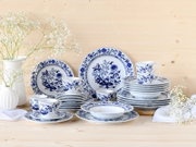 Blau-weißes Porzellan-Geschirrset mit floralem Zwiebelmuster, gestapelte Teller, Schalen und Tassen auf hellem Holztisch neben weißen Vasen mit Schleierkraut, Frontansicht auf Augenhöhe.