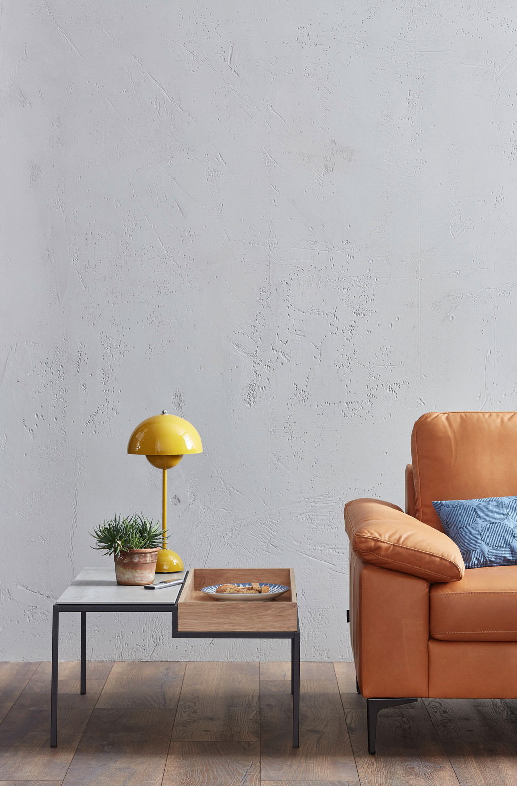 Seitliche Ansicht eines modernen 2,5-Sitzer Sofas in hellbraunem Leder, neben einem Couchtisch mit gelber Lampe und Pflanze.