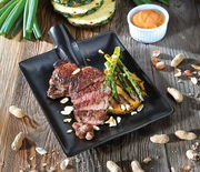 Schwarzer Steak-Servierteller mit gegrilltem Steak, Spargel und Ananas, dekoriert mit Nüssen, aus der Vogelperspektive.