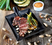 Schwarzer Steak-Servierteller mit gegrilltem Steak, Spargel und Ananas, dekoriert mit Nüssen, aus der Vogelperspektive.