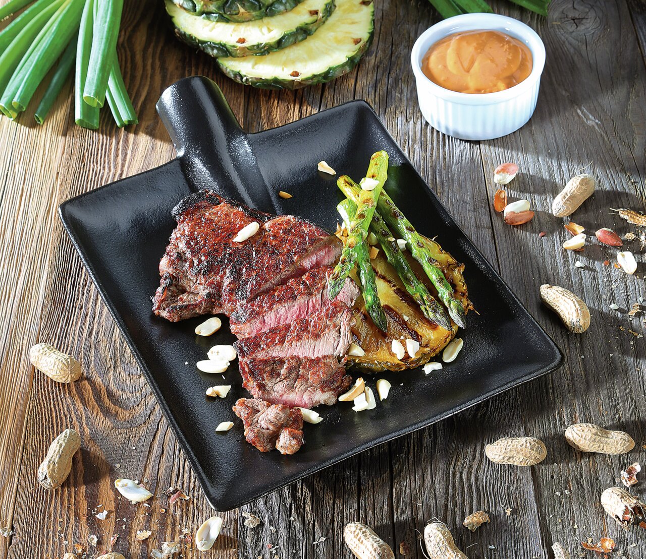 Zassenhaus Steak-/serveerbord WORKER Zwart serveerbord met een gegrilde steak, asperges en ananas, gegarneerd met noten, gezien vanuit vogelperspectief.