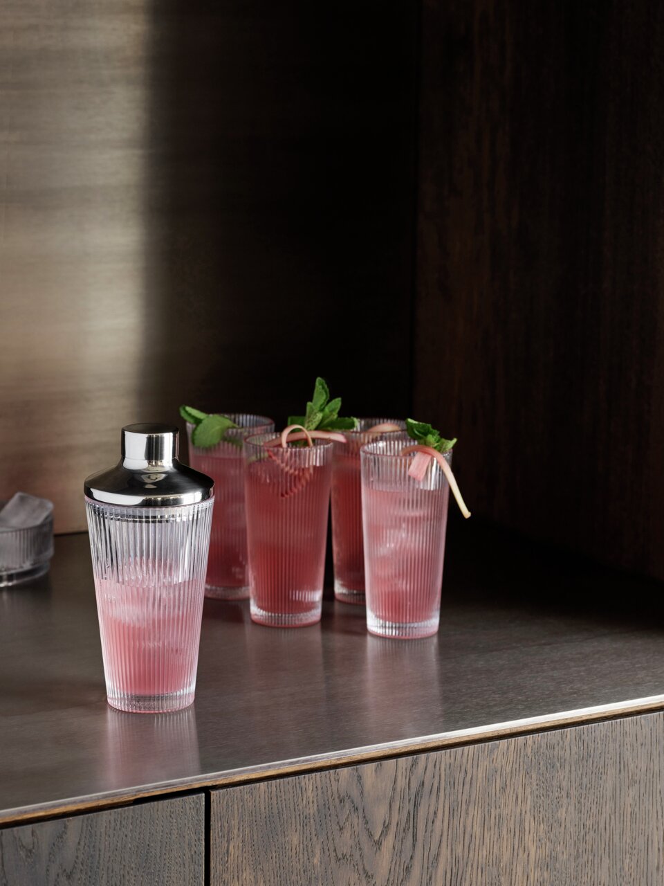 Stelton Cocktailshaker PILASTRO Pilastro Cocktail Shaker aus klarem Glas mit geriffeltem Design, auf einer Arbeitsplatte stehend, umgeben von gefüllten Gläsern mit rosa Getränken, seitliche Perspektive.