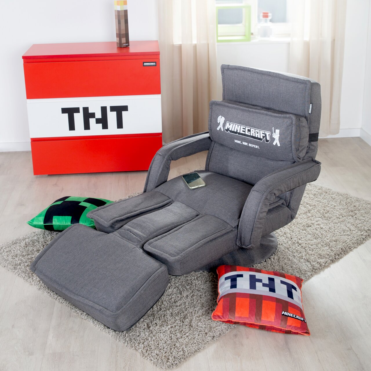 Grauer Minecraft-Gaming-Sessel mit Armlehnen, hoher Rückenlehne und ausklappbarer Fußstütze auf Teppich, schräg von vorne rechts fotografiert.