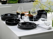 Schwarzes, glänzendes Geschirr-Set mit Tellern, Schalen und Besteck auf weißem Tisch, dekoriert mit Weingläsern und Blumen, aus leicht schräger Seitenperspektive