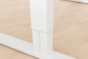 Close-up van een witte, in hoogte verstelbare tafelpoot met hoogteaanduidingen op een lichte houten vloer.