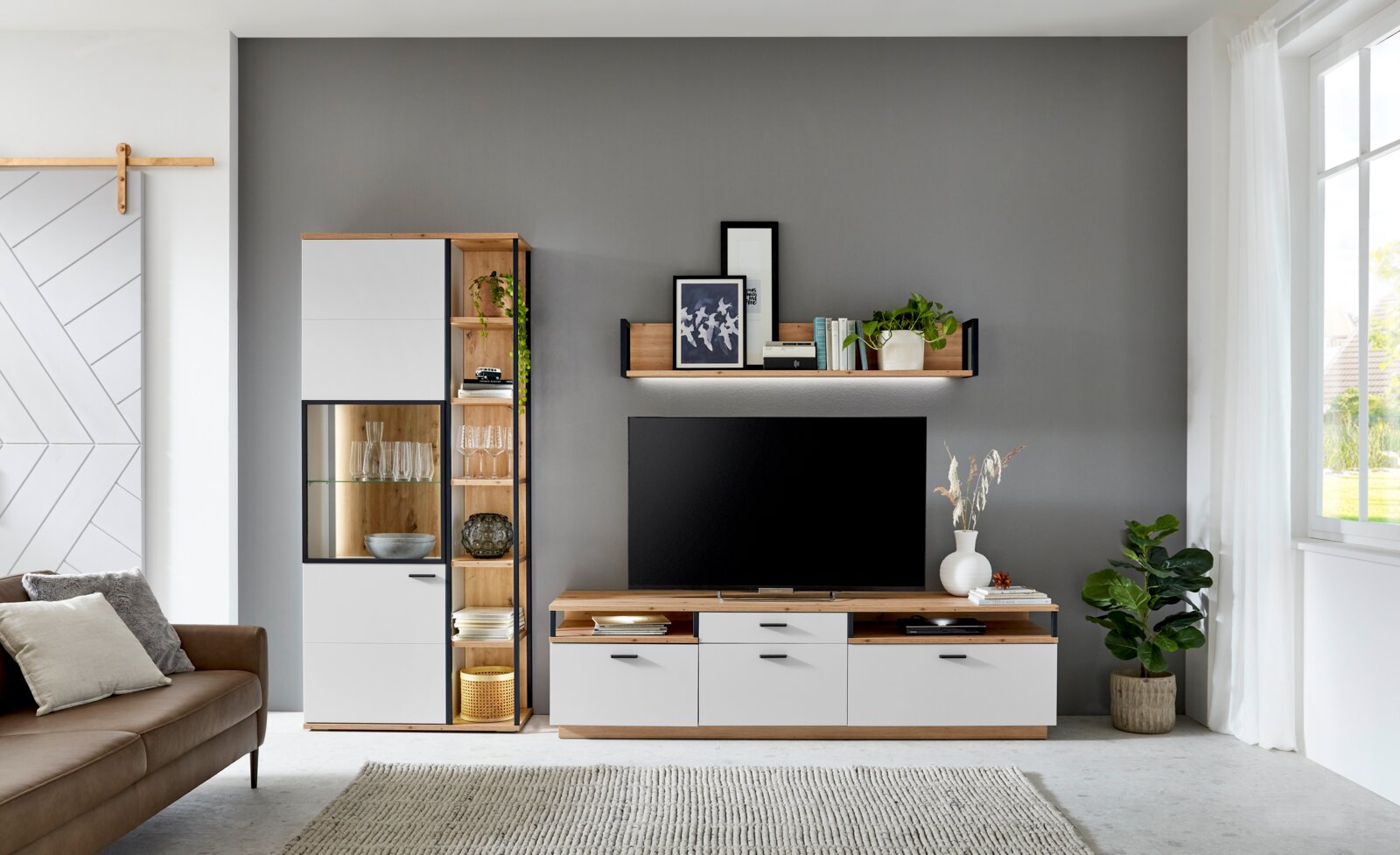 Moderne Wohnzimmer-Wandkombination in Holzoptik mit weißen Fronten, bestehend aus einem hohen Schrank, einem TV-Board und einem Wandregal, frontal fotografiert.