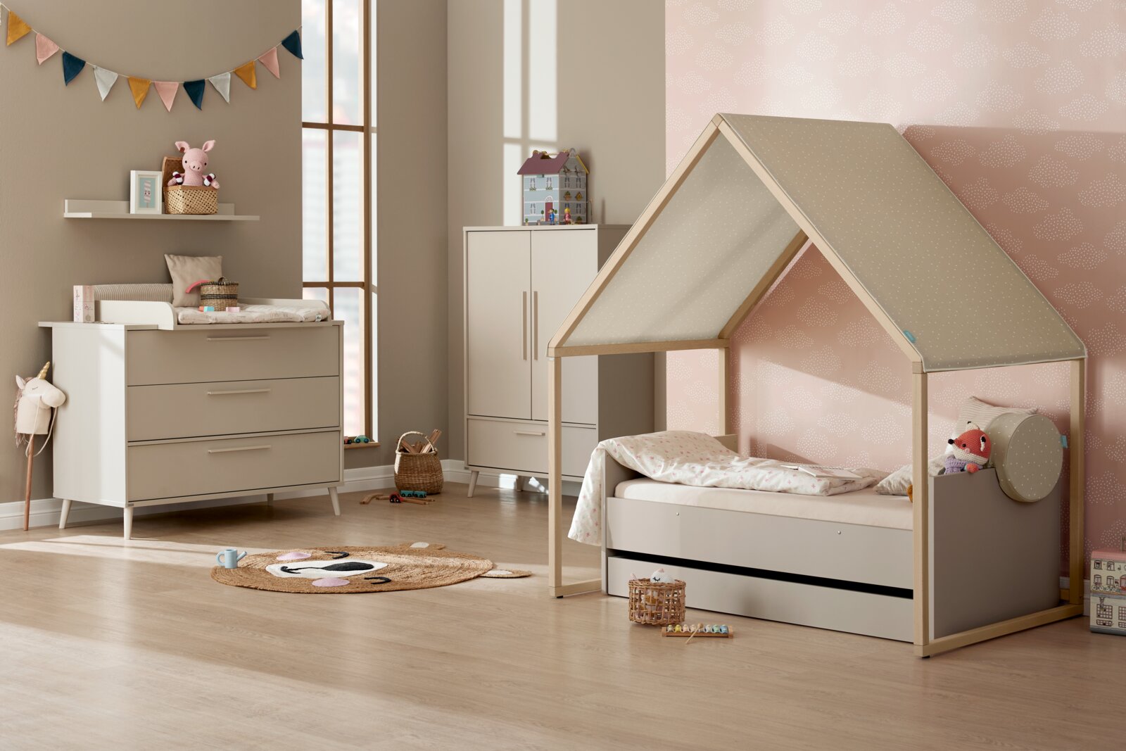 Gemütliches Kinderzimmer mit Hausbett in Naturholz und grauem Stoffdach, aufgenommen aus seitlicher Frontperspektive; daneben Kommode und Schrank in Beige-Weiß sowie Dekorationen.