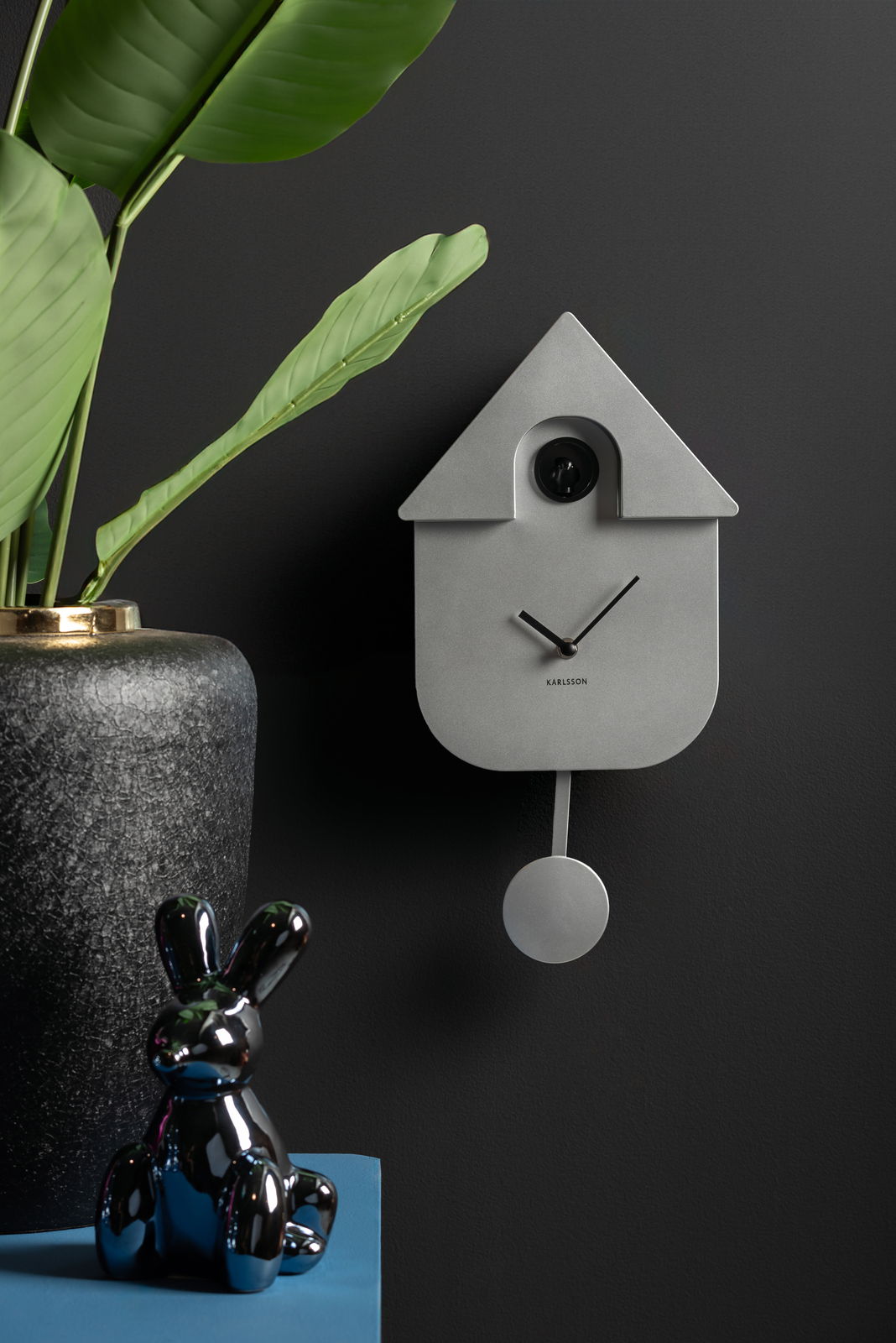 Moderne Wanduhr in Form eines minimalistischen Kuckucksuhren-Designs, frontal an einer dunklen Wand montiert, mit einem Pendel und umgeben von dekorativen Elementen.