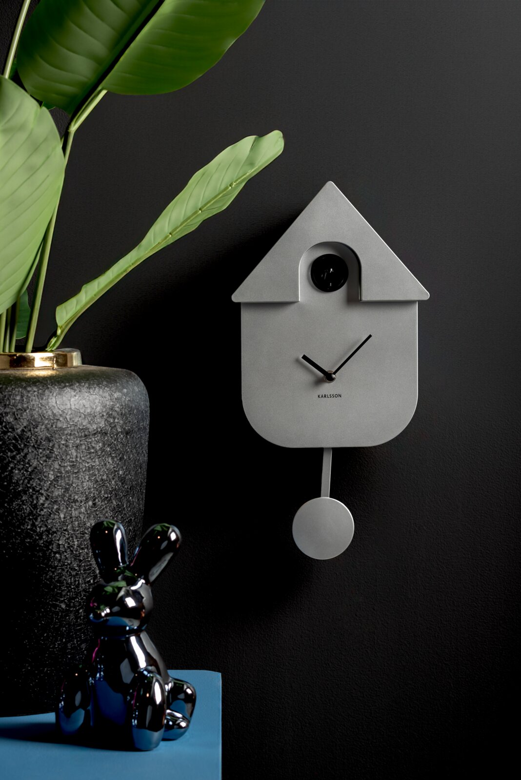 Moderne Wanduhr in Form eines minimalistischen Kuckucksuhren-Designs, frontal an einer dunklen Wand montiert, mit einem Pendel und umgeben von dekorativen Elementen.