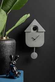Moderne Wanduhr in Form eines minimalistischen Kuckucksuhren-Designs, frontal an einer dunklen Wand montiert, mit einem Pendel und umgeben von dekorativen Elementen.