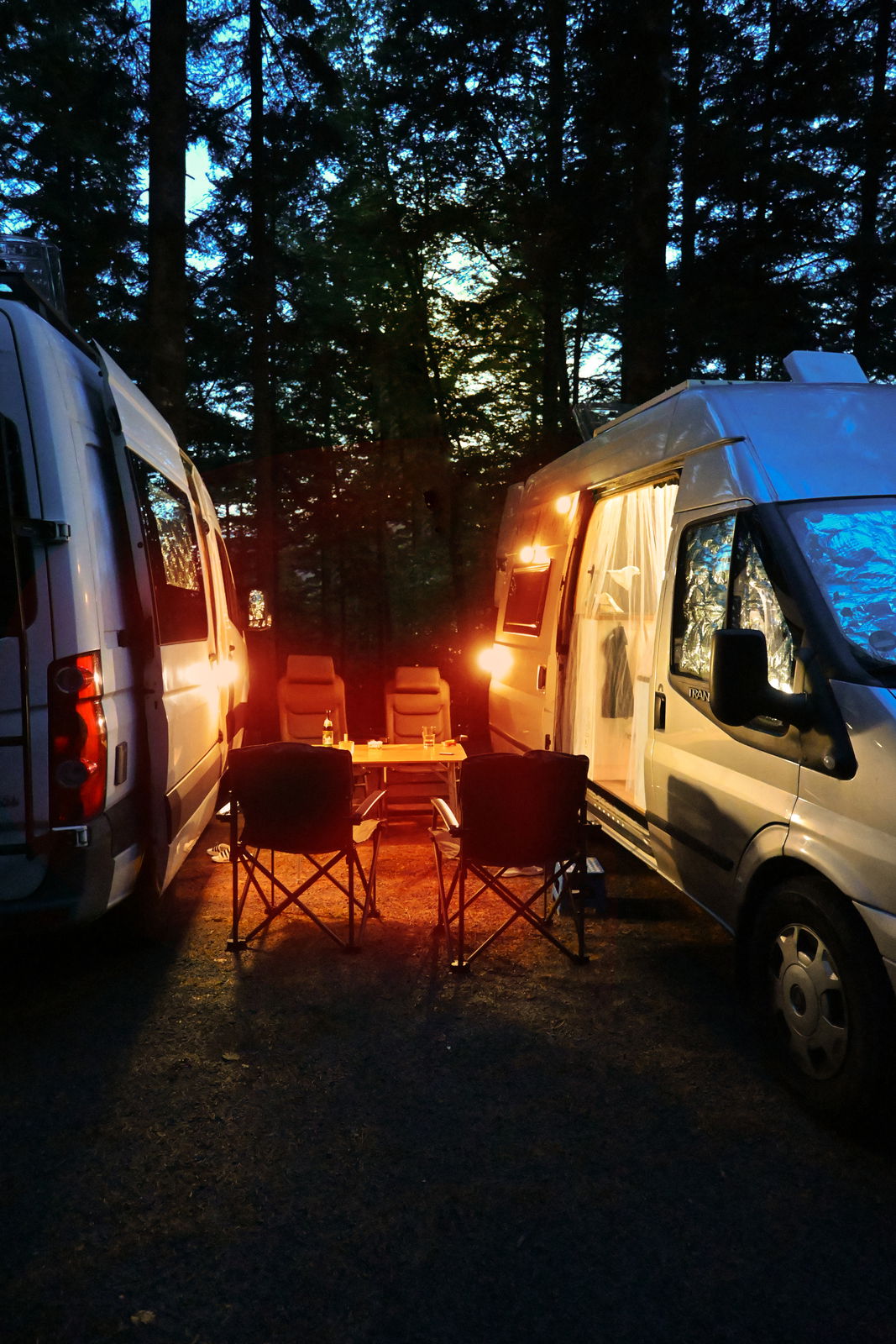 Camping-Szene bei Dämmerung mit zwei Wohnmobilen, die ein gemütliches Licht auf einen Tisch und Stühle werfen, seitliche Perspektive.
