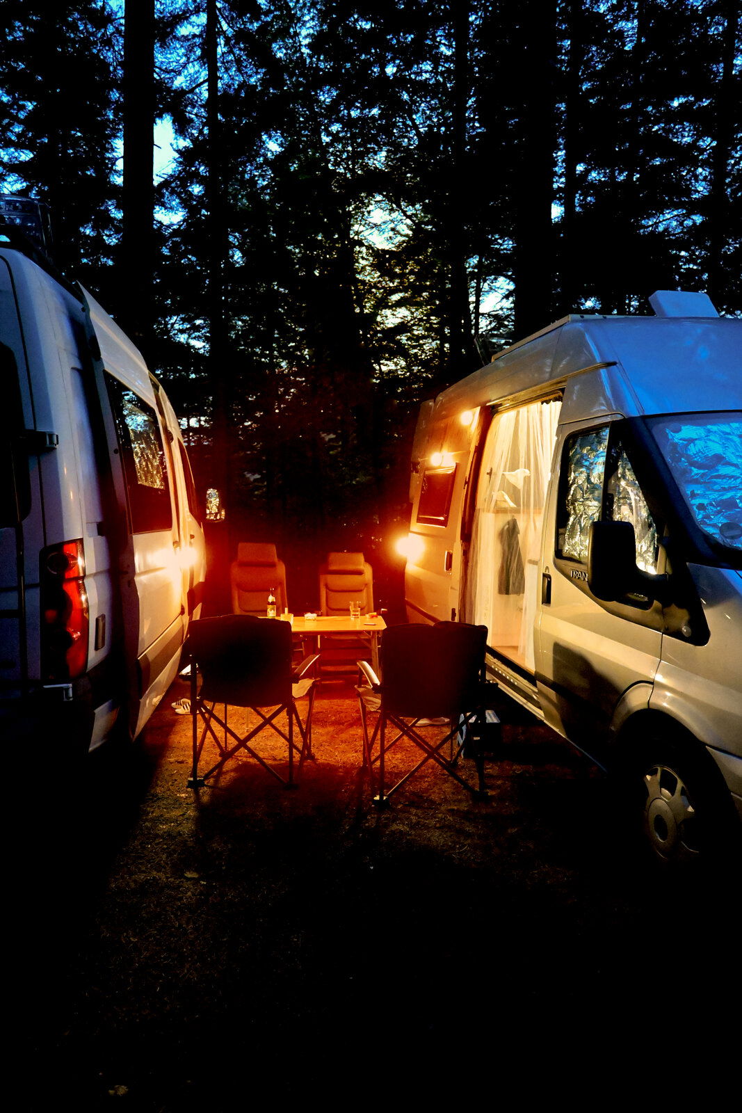 Camping-Szene bei Dämmerung mit zwei Wohnmobilen, die ein gemütliches Licht auf einen Tisch und Stühle werfen, seitliche Perspektive.