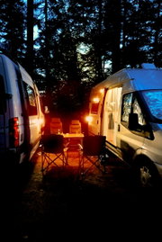 Camping-Szene bei Dämmerung mit zwei Wohnmobilen, die ein gemütliches Licht auf einen Tisch und Stühle werfen, seitliche Perspektive.