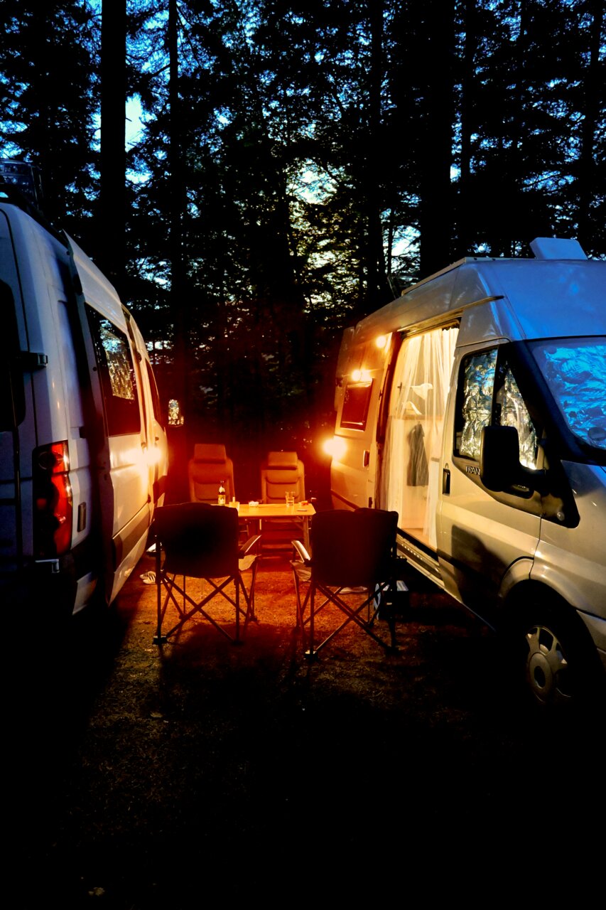 Campingtafereel bij schemering met twee campers die een gezellig licht op een tafel en stoelen werpen, zijaanzicht.