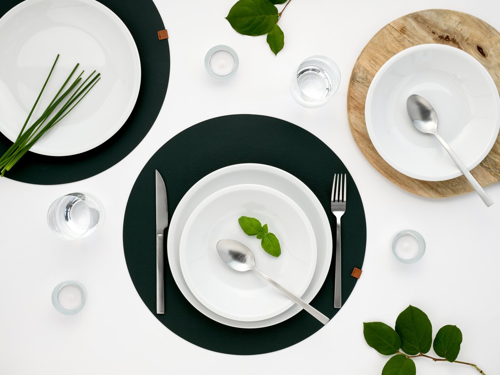 Draufsicht auf ein modern gedecktes Tisch-Arrangement mit weißen Porzellantellern und Schalen auf dunklen Platzsets, Besteck und Wassergläsern, ergänzt durch grüne Kräuter und Blätter auf weißem Hintergrund