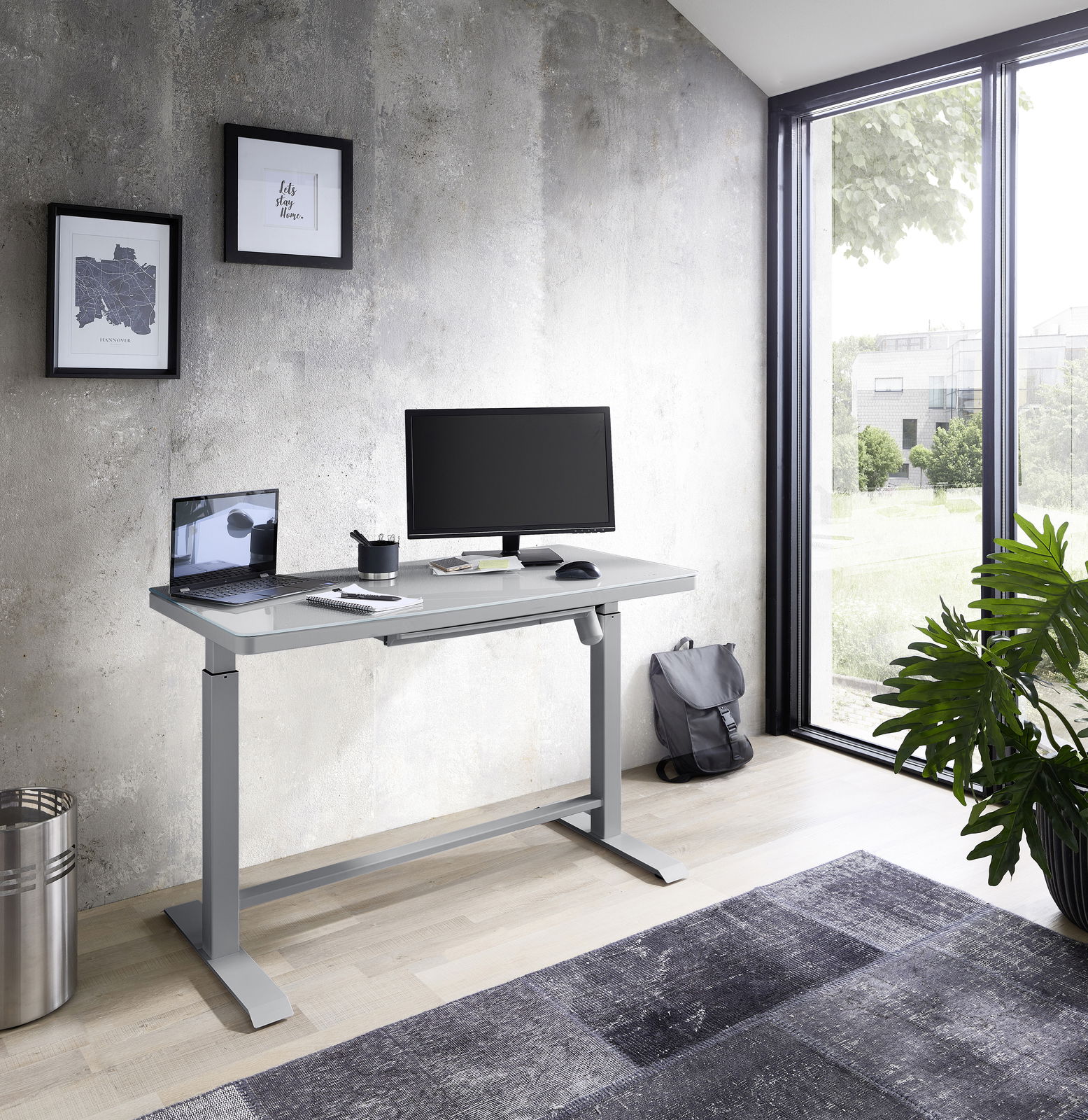 Moderner höhenverstellbarer E-Schreibtisch in einem hellen Raum mit Blick auf ein großes Fenster, ausgestattet mit einem Monitor, Laptop und Schreibtischzubehör.