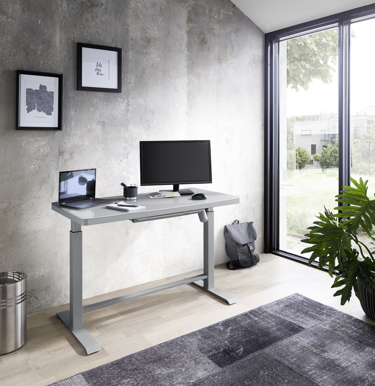 Moderner höhenverstellbarer E-Schreibtisch in einem hellen Raum mit Blick auf ein großes Fenster, ausgestattet mit einem Monitor, Laptop und Schreibtischzubehör.
