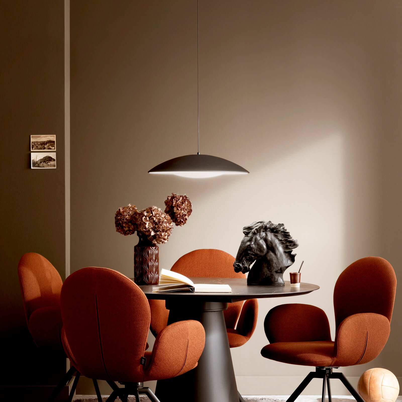 Modernes Esszimmer-Setting mit schwarzer, flacher Pendelleuchte über rundem Tisch, orangefarbenen Polsterstühlen und Deko (Vase mit Trockenblumen, Pferdekopf-Skulptur), aufgenommen in frontaler Perspektive auf Augenhöhe.