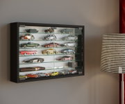 Schwarze Wandvitrine mit Glasfront und mehreren Glasböden, gefüllt mit Modellautos, in schräger Frontansicht von links an einer grauen Wand neben einer Stehlampe.