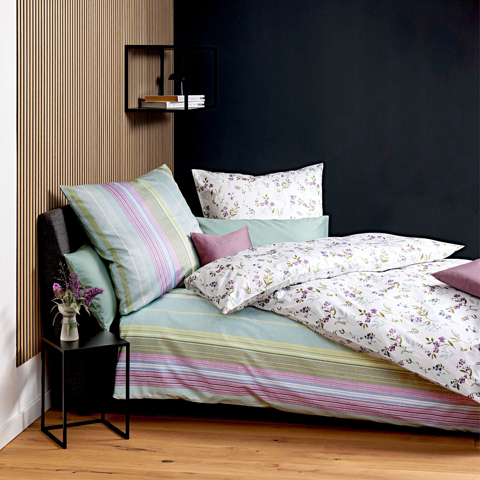 Modernes Bett mit weißer Bettwäsche mit zartem Blumenmuster und gestreiftem Spannbettlaken in Pastellfarben, dekoriert mit Kissen; Ansicht schräg von vorne aus leicht erhöhter Perspektive.