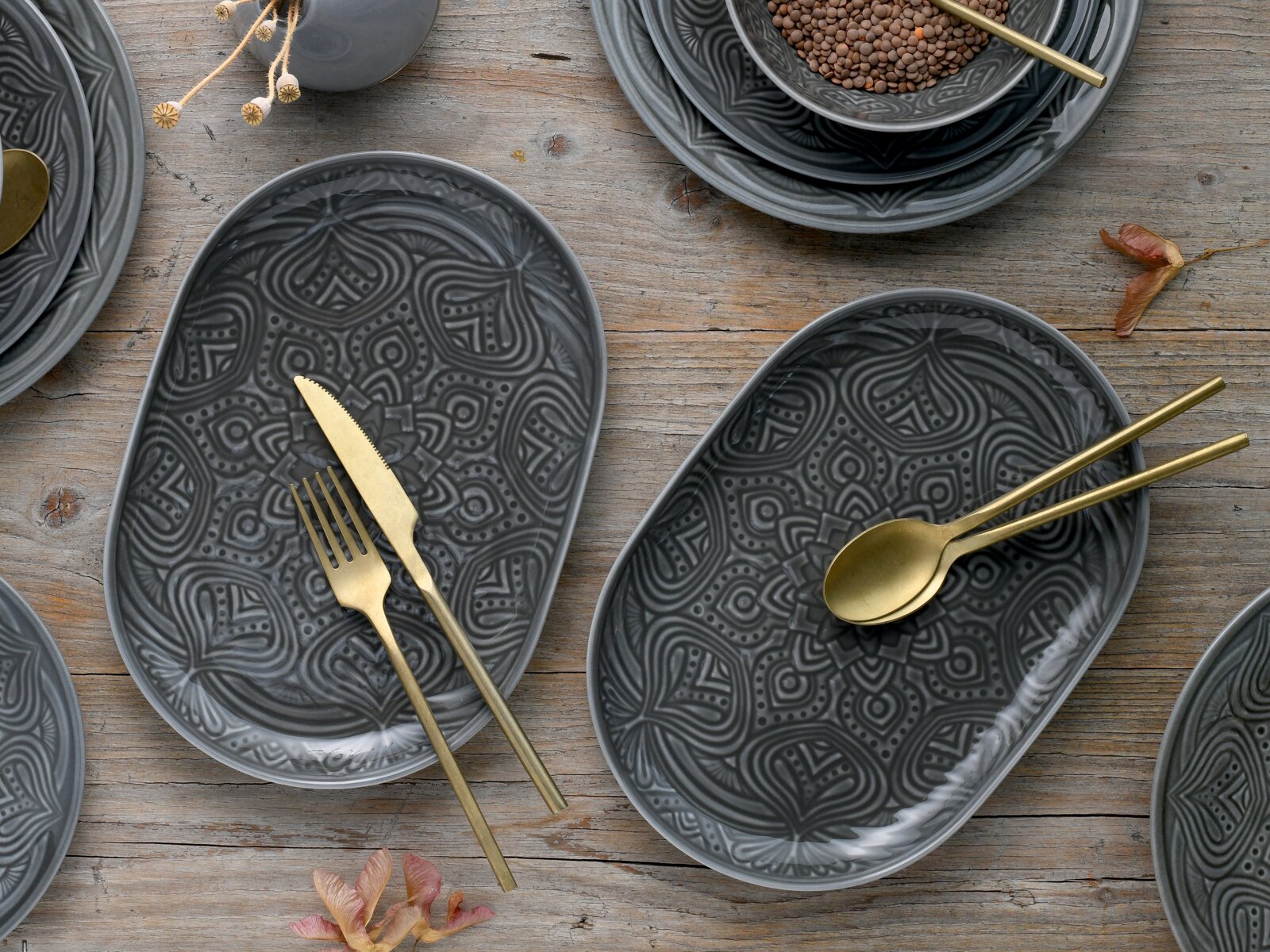 Dunkelgraue, ovale Keramik-Servierplatten mit ornamentalem Reliefmuster auf rustikalem Holztisch, dekoriert mit goldfarbenem Besteck; Draufsicht von oben