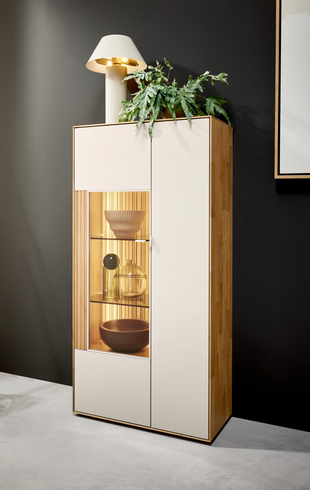 Hoher Vitrinenschrank aus hellem Holz mit weißen Fronten und beleuchtetem Glasfach, dekoriert mit Schalen und Vasen, aufgenommen in leichter Schrägansicht von vorne