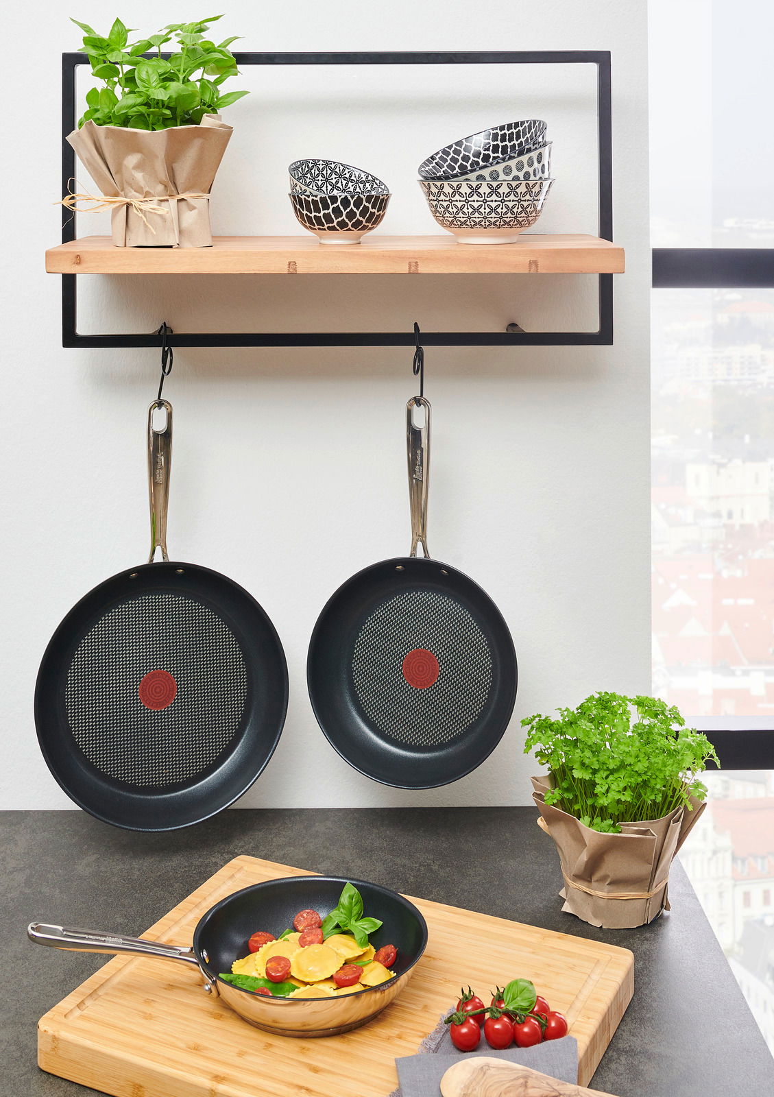 Drei Bratpfannen Jamie Oliver Smart, zwei hängend und eine auf einem Schneidebrett mit Ravioli und Tomaten, aus der Perspektive von vorne.