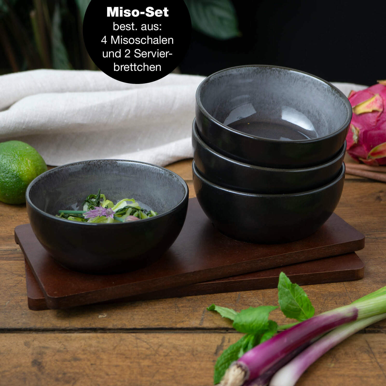 Vier anthrazitfarbene Miso-Schalen, gestapelt auf zwei Servierbrettern, mit einer Schale im Vordergrund, gefüllt mit frischen Kräutern, aus seitlicher Perspektive.