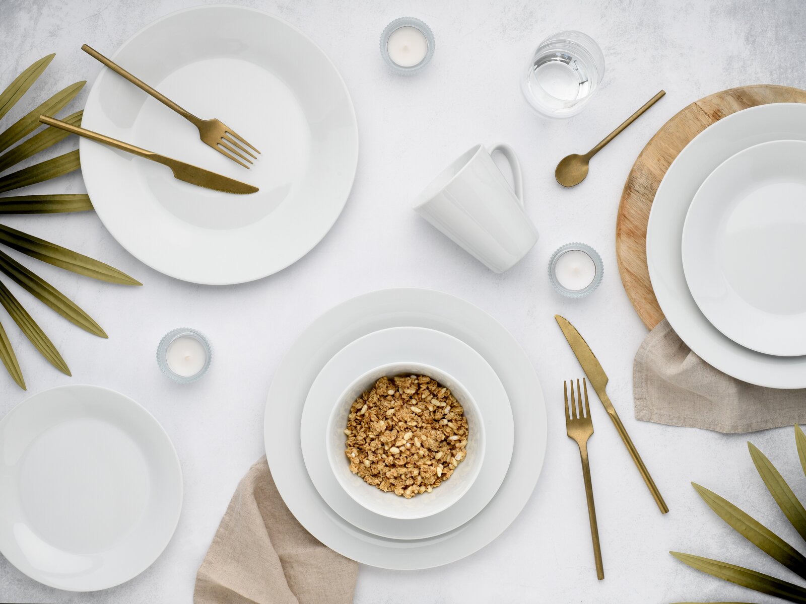 Draufsicht auf ein modern gedecktes Tischarrangement mit weißen Tellern und Schalen, goldfarbenem Besteck, weißer Tasse, Wasserglas, kleinen Teelichtern und einer Schale Müsli auf hellem Hintergrund
