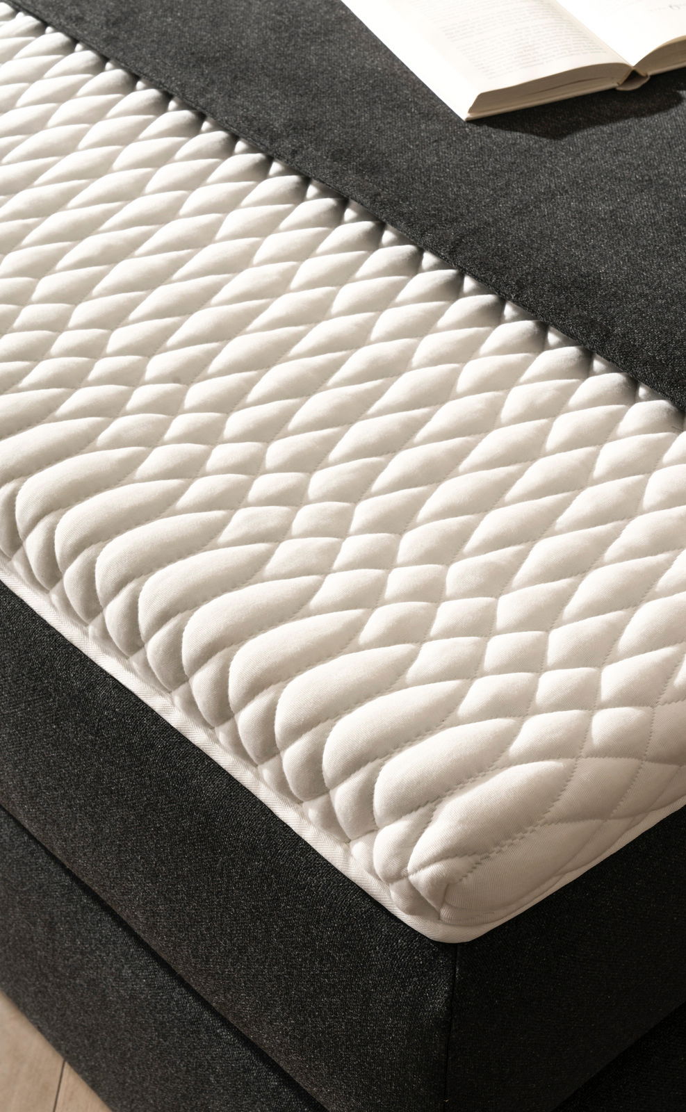 Nahaufnahme eines Boxspringbetts mit weißer, gesteppter Matratzenauflage und dunklem Stoffbezug, von schräg oben fotografiert.