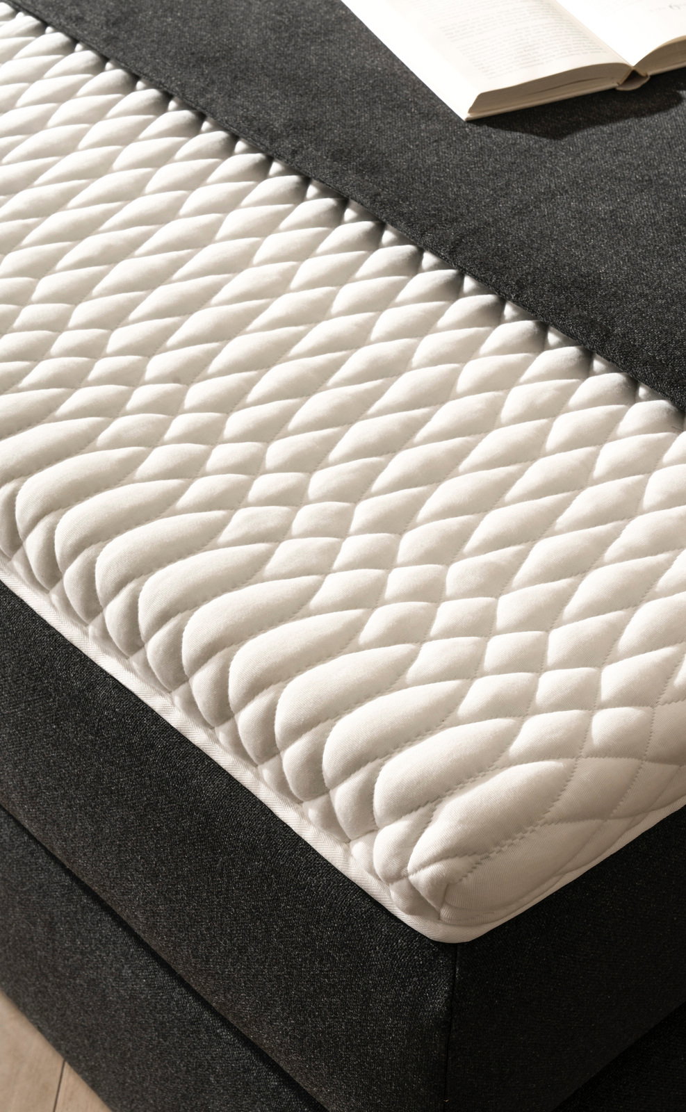 Nahaufnahme eines Boxspringbetts mit weißer, gesteppter Matratzenauflage und dunklem Stoffbezug, von schräg oben fotografiert.