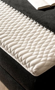 Nahaufnahme eines Boxspringbetts mit weißer, gesteppter Matratzenauflage und dunklem Stoffbezug, von schräg oben fotografiert.