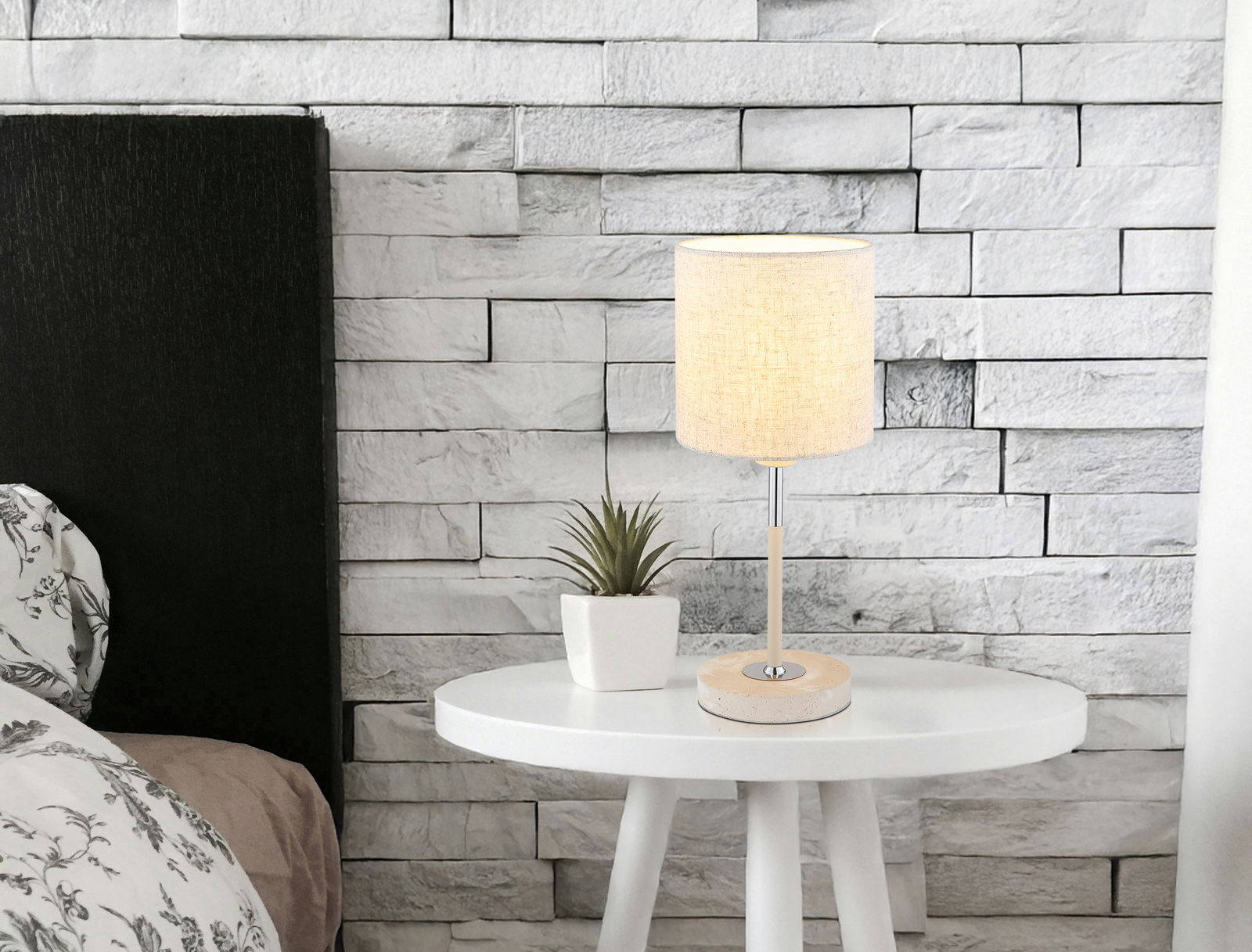 Tischleuchte auf einem weißen runden Tisch, neben einer kleinen Pflanze, vor einer weißen Ziegelwand. Perspektive: seitlich.