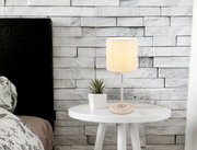 Tischleuchte auf einem weißen runden Tisch, neben einer kleinen Pflanze, vor einer weißen Ziegelwand. Perspektive: seitlich.