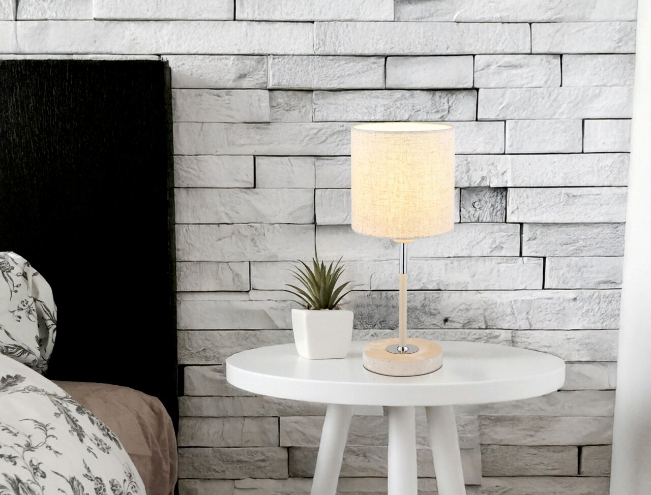 Tischleuchte auf einem weißen runden Tisch, neben einer kleinen Pflanze, vor einer weißen Ziegelwand. Perspektive: seitlich.