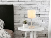 Tischleuchte auf einem weißen runden Tisch, neben einer kleinen Pflanze, vor einer weißen Ziegelwand. Perspektive: seitlich.