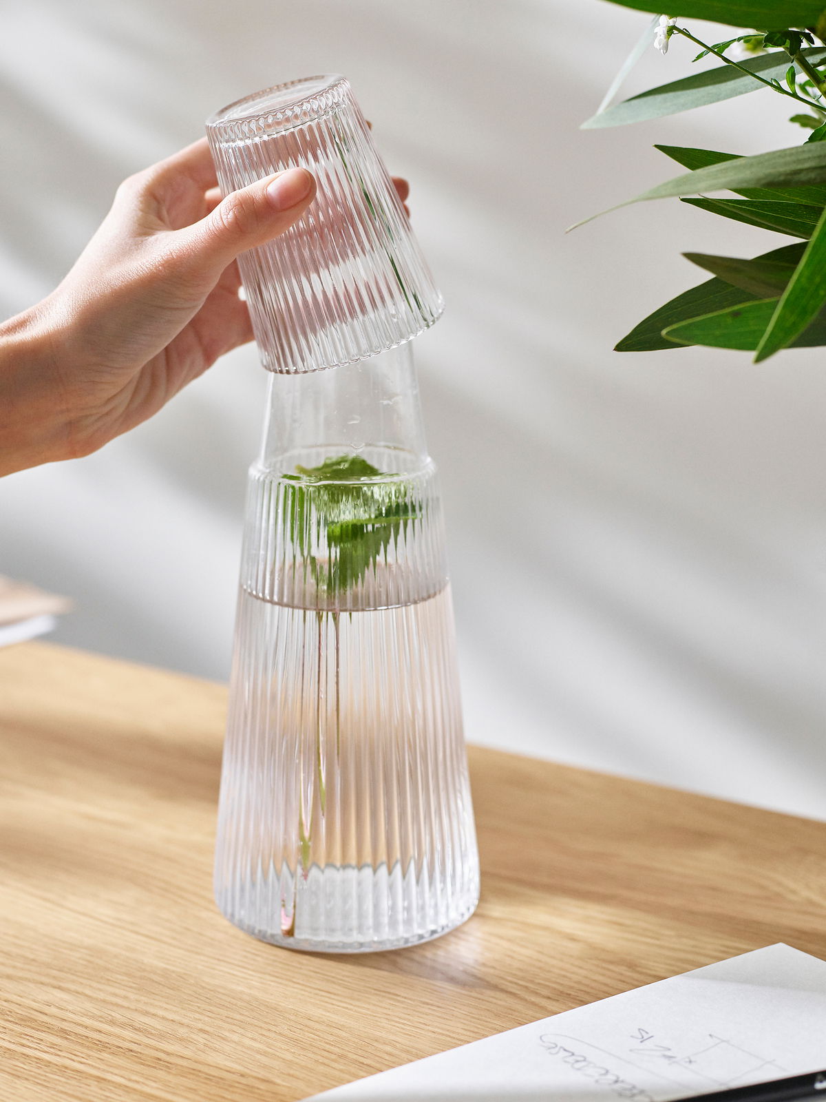 Eine Hand hebt ein geriffeltes Glas von einer Pilastro Karaffe mit Wasser und einem Minzzweig, auf einem Holztisch stehend, ab. Seitliche Perspektive.