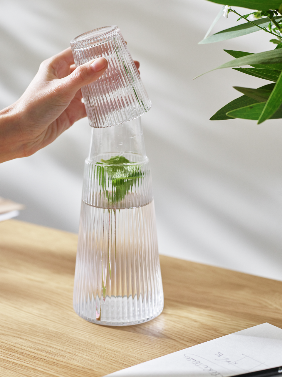 Eine Hand hebt ein geriffeltes Glas von einer Pilastro Karaffe mit Wasser und einem Minzzweig, auf einem Holztisch stehend, ab. Seitliche Perspektive.